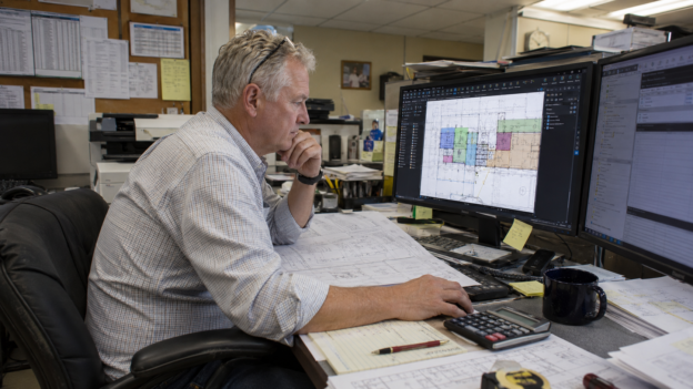 Senior estimator at a dual-monitor workstation with Bluebeam Revu open on a takeoff, printed plans nearby.