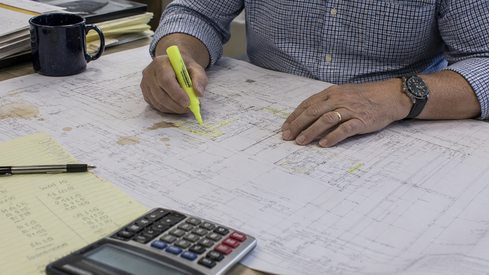 Senior estimator marking up printed construction plans by hand with a highlighter and calculator.